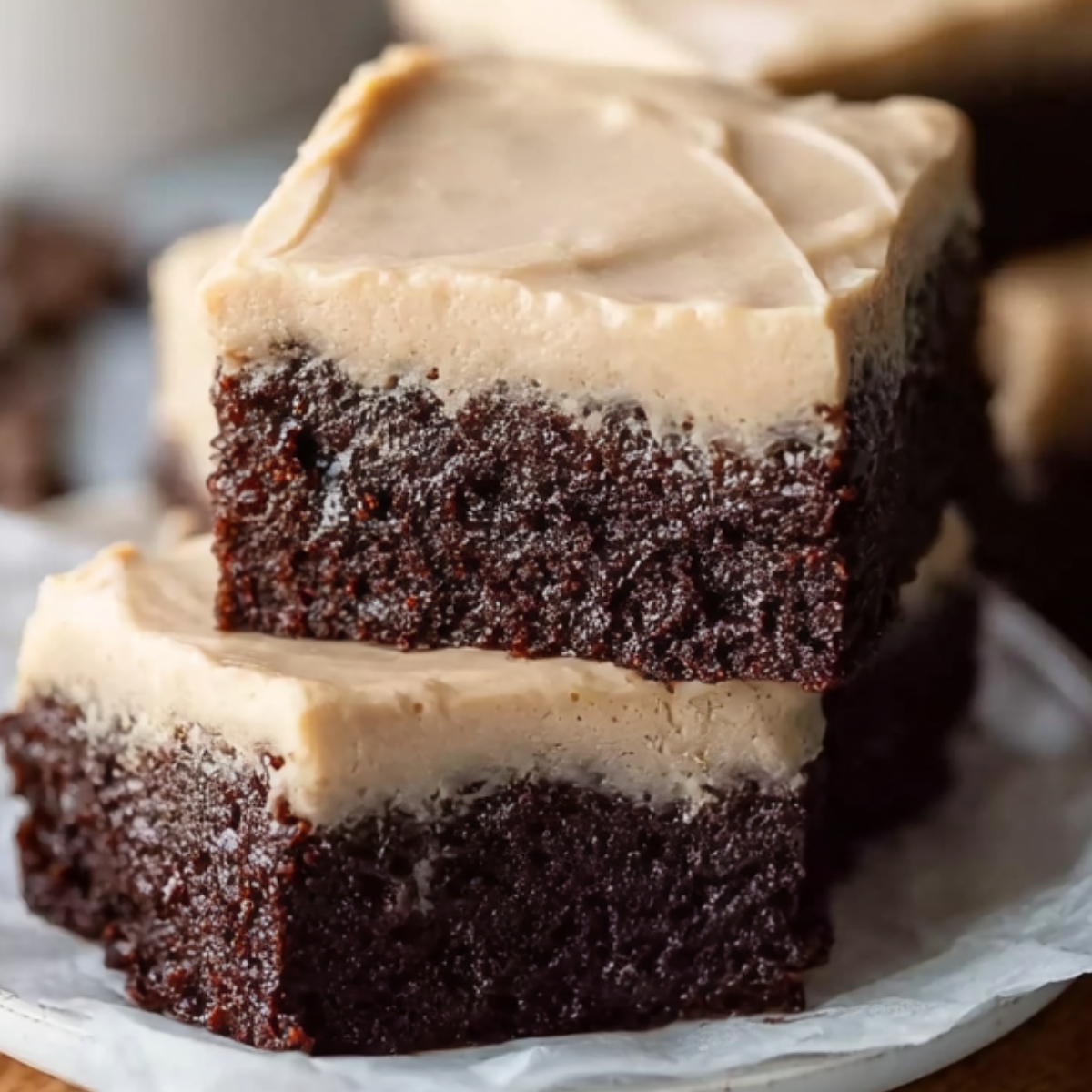 Frosted coffee brownies on a plate, showcasing dense chocolate layers topped with creamy coffee frosting, with a cup of coffee in the background.