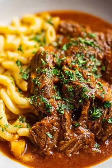 A plate of hearty German Goulash with thick pasta noodles, coated in rich reddish-brown sauce and sprinkled with fresh parsley.