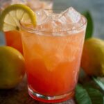 Guava White Tea Lemonade Two glasses of iced pink guava lemonade, each with a lemon slice on the rim. Whole guavas and green leaves sit around them on a textured surface.