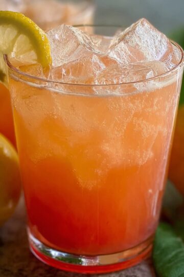 Guava White Tea Lemonade Two glasses of iced pink guava lemonade, each with a lemon slice on the rim. Whole guavas and green leaves sit around them on a textured surface.
