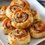 Freshly baked ham and cheese pinwheels on a white serving plate, flaky and golden with melted cheese and herbs