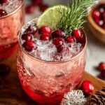 Holiday Mule Cocktails “Two textured copper mugs with a red layered drink and crushed ice, topped with cranberries, a lime wedge, and rosemary. Cranberries and holiday décor appear in the background.