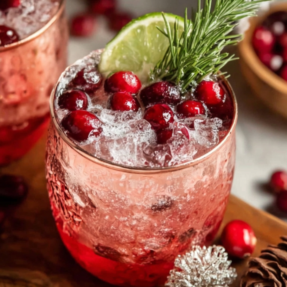Holiday Mule Cocktails “Two textured copper mugs with a red layered drink and crushed ice, topped with cranberries, a lime wedge, and rosemary. Cranberries and holiday décor appear in the background.