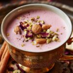 A decorative teacup filled with frothy pink Kashmiri chai, topped with crushed pistachios and dried rose petals, placed on an ornate metal tray with cinnamon sticks and scattered rose petals.