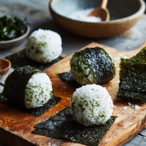 Small rice balls wrapped with strips of seaweed arranged on a wooden board, with extra nori sheets and salt in the background.