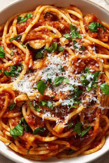 Pasta Alla Norma A plate of thick spaghetti coated in rich tomato sauce, sprinkled generously with grated cheese and fresh basil, with a glass of white wine beside it.