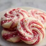 Peppermint Swirl Cookies A plate of peppermint swirl cookies coated in sugar, with red and white spiral centers and crushed peppermint pieces sprinkled on top.