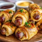 Pigs In A Blanket A plate of golden, flaky pigs in a blanket made with puff pastry, stacked neatly and garnished with parsley, served with a small dish of mustard.