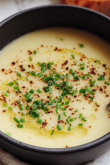 A smooth potato leek soup served in a black ceramic bowl, garnished with finely chopped chives, olive oil drizzle, and cracked pepper, with crusty bread and a spoon beside it on a light cloth.
