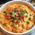 A serving of creamy shredded Queso Chicken Crockpot in a bowl, garnished with chopped tomatoes, green onions, and melted cheese.