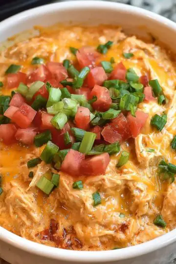 A serving of creamy shredded Queso Chicken Crockpot in a bowl, garnished with chopped tomatoes, green onions, and melted cheese.