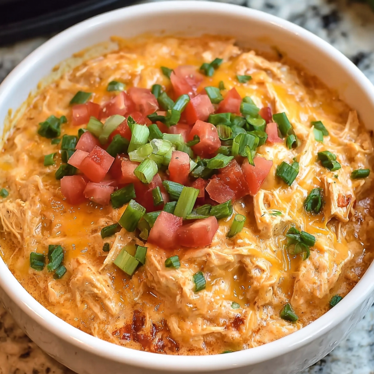 A serving of creamy shredded Queso Chicken Crockpot in a bowl, garnished with chopped tomatoes, green onions, and melted cheese.