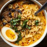 Spicy Coconut Curry Ramen A bowl of creamy orange coconut curry ramen topped with sautéed mushrooms, fresh cilantro, sesame seeds, chili flakes, and half a soft-boiled egg.