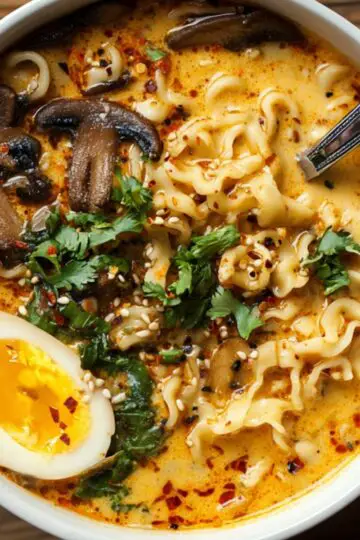 Spicy Coconut Curry Ramen A bowl of creamy orange coconut curry ramen topped with sautéed mushrooms, fresh cilantro, sesame seeds, chili flakes, and half a soft-boiled egg.
