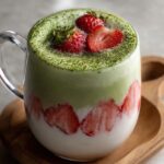 A creamy strawberry matcha latte served in a glass mug on a wooden tray, with layers of pink strawberry milk, white cream, and green matcha, topped with fresh strawberries and matcha powder.
