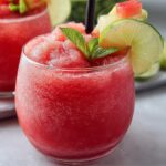 Watermelon Slushie Recipe Two round glasses filled with bright red watermelon slushie, garnished with a small watermelon wedge and mint leaves, sitting on a plate with ice."