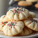 Close-up of tender whipped shortbread cookies topped with red, green, and white holiday sprinkles, showing their delicate, crumbly texture on a decorative plate."
