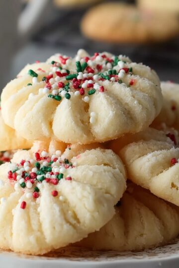 Close-up of tender whipped shortbread cookies topped with red, green, and white holiday sprinkles, showing their delicate, crumbly texture on a decorative plate."