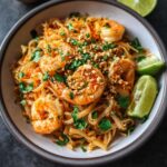 A plate of shrimp Pad Thai topped with chopped green onions and crushed peanuts, served with lime wedges and fresh cilantro.