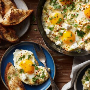 The Best Cottage Cheese Eggs Recipe 17 A rustic breakfast spread featuring a skillet filled with sunny eggs topped with herbs and cottage cheese, surrounded by toasted bread, avocado chunks, and plates with servings of the egg dish.