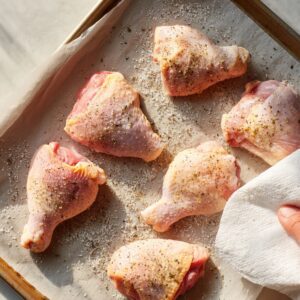 Delicious Baked Lemon Butter Chicken Recipe 15 Raw chicken pieces on a parchment-lined baking sheet being patted dry with a paper towel, seasoned with salt, pepper, and herbs, ready for roasting.