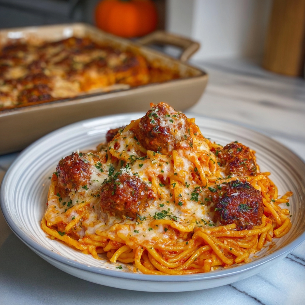 A plate of baked spaghetti topped with meatballs and melted cheese, garnished with herbs, with a casserole dish of more pasta in the background.
