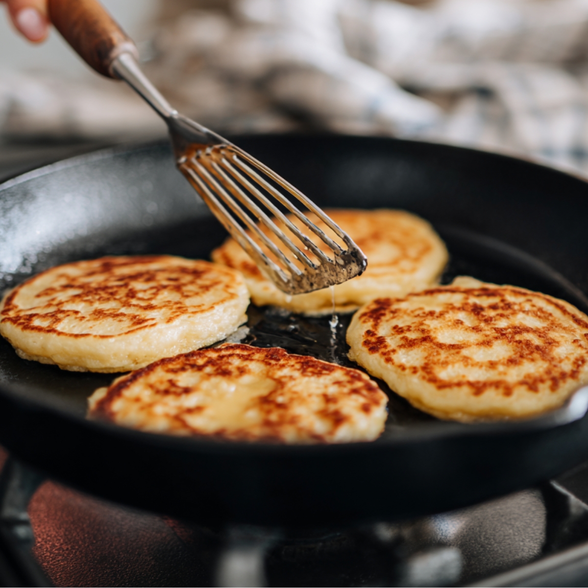 Healthy Banana Pancakes Recipe 12 A close-up of four golden-brown pancakes sizzling in a black cast-iron skillet on a stovetop. A hand holding a metal slotted spatula gently lifts one pancake, showing its crisp, evenly browned surface. The background is softly blurred, with a striped kitchen towel adding a cozy, home-cooking atmosphere.