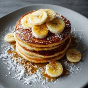 Healthy Banana Pancakes Recipe 19 A tall stack of fluffy, golden-brown banana pancakes sits on a gray plate. Thin slices of banana are arranged on top, drizzled with glossy syrup. A dusting of shredded coconut surrounds the stack and melts slightly into the syrup. The lighting enhances the rich texture and appetizing shine of the pancakes.