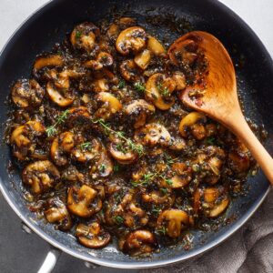 Best Beef Wellington Bites Recipe 15 A skillet filled with deeply caramelized mushrooms, shallots, garlic, and thyme—classic mushroom duxelles—stirred with a wooden spoon.