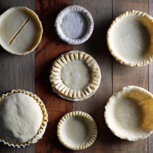 The Best British Meat Pies Recipe 17 Several unbaked pie shells and pastry tops arranged on a wooden surface, showing different sizes and crimped edge designs before filling and sealing.