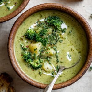 A bowl of creamy broccoli and potato soup topped with broccoli florets, herbs, and a drizzle of cream, served with bread on the side.