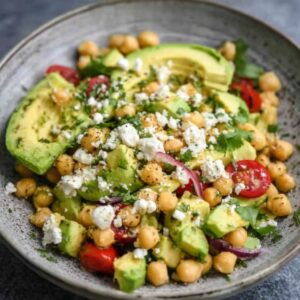 A plate featuring sliced avocado, chickpeas, cherry tomatoes, red onion, crumbled feta cheese, and fresh herbs.
