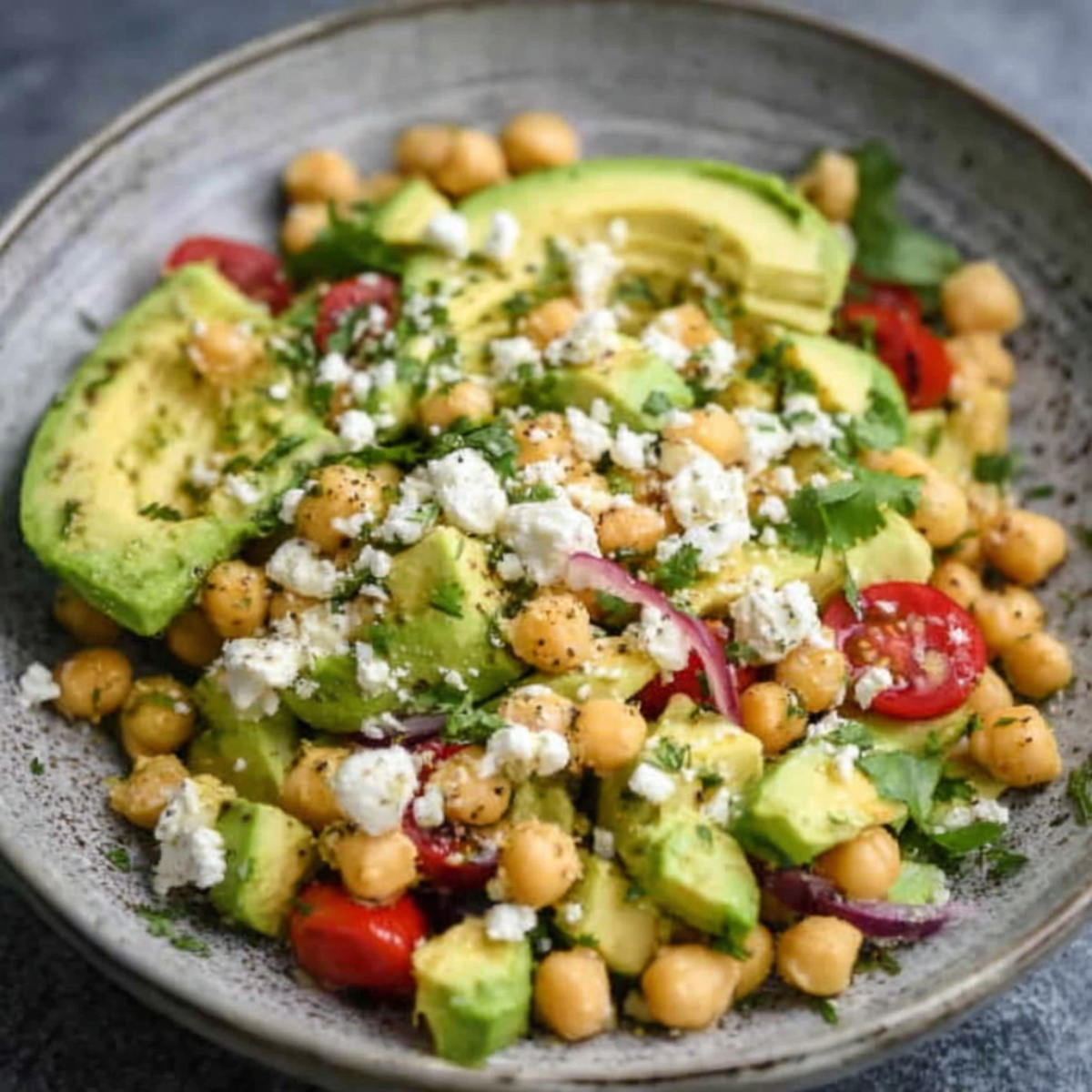 A plate featuring sliced avocado, chickpeas, cherry tomatoes, red onion, crumbled feta cheese, and fresh herbs.