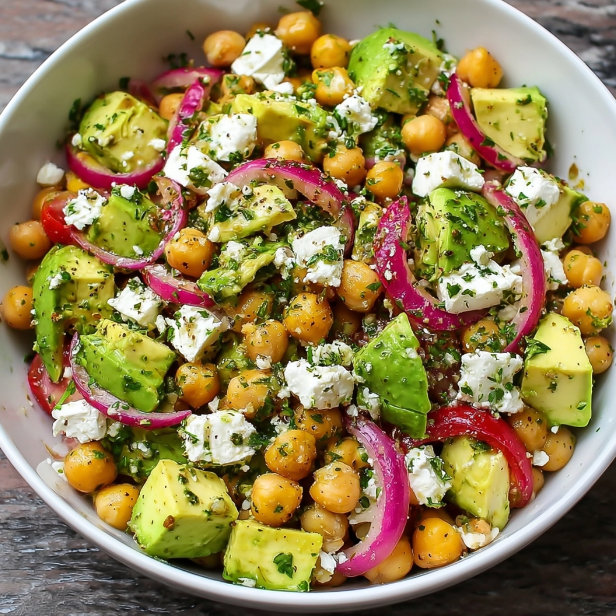 A chickpea salad with avocado chunks, red onion slices, cherry tomatoes, and crumbled feta, mixed with herbs and dressing in a white bowl.