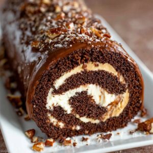 A Chocolate Cake Roll roll filled with creamy vanilla frosting, topped with chocolate drizzle, chopped nuts, and a dusting of powdered sugar on a white serving plate.