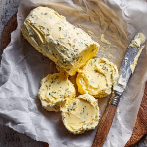 Rolled and sliced homemade herb butter with visible green herbs, placed on parchment paper with a knife beside it.