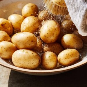 Easy Slow Cooker Lipton Onion Potatoes Recipe 13 Hands scrubbing baby potatoes under running water using a vegetable brush over a bowl. Water splashes as dirt is removed, with a kitchen towel nearby. Clean, freshly washed potato skins glisten in natural light.