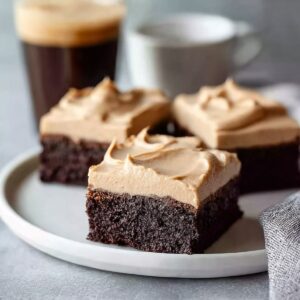 Close-up of frosted coffee brownies on a white plate, highlighting the thick, smooth layer of coffee frosting over moist chocolate brownie squares.