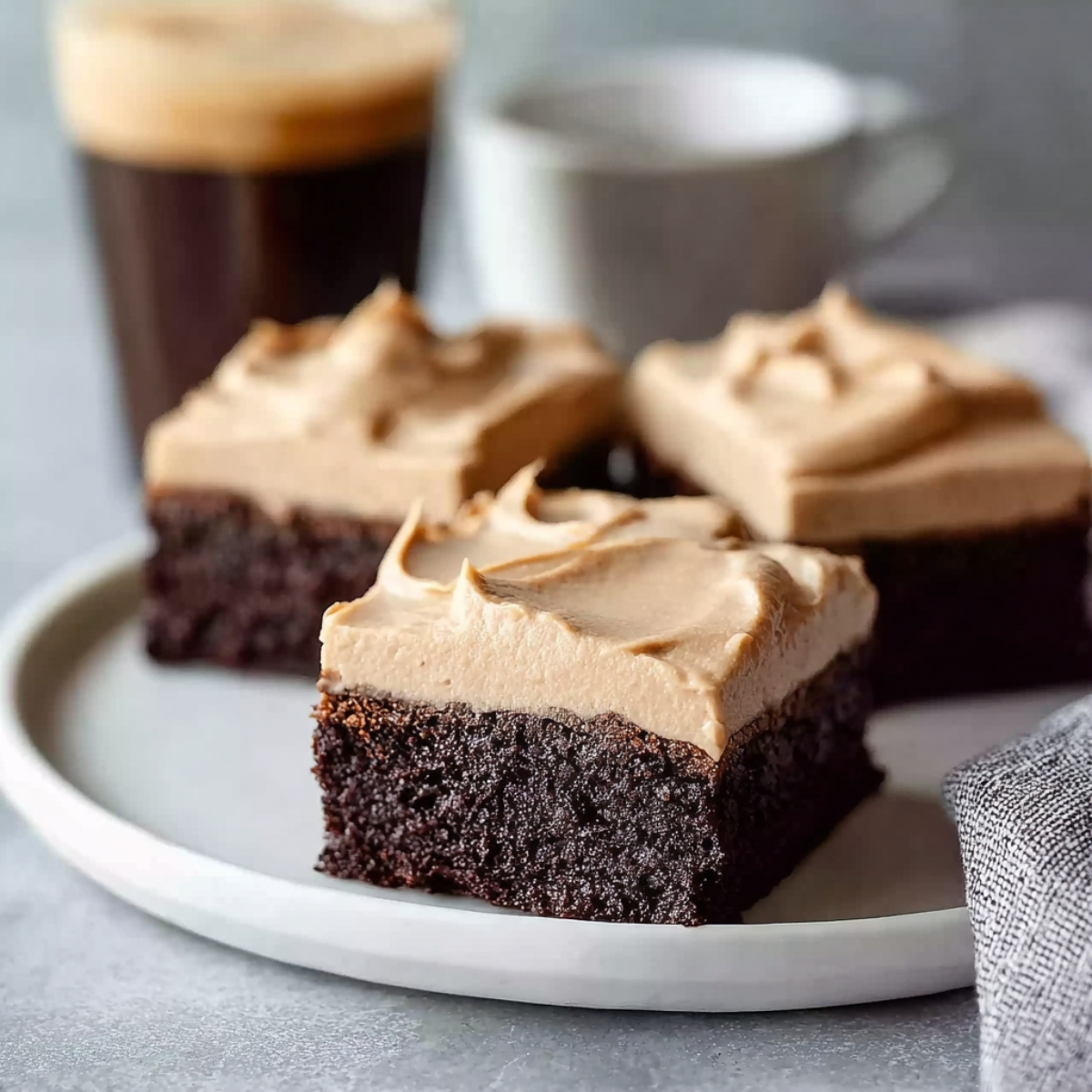 Close-up of frosted coffee brownies on a white plate, highlighting the thick, smooth layer of coffee frosting over moist chocolate brownie squares.