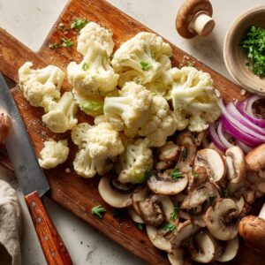 A wooden cutting board covered with freshly chopped cauliflower florets and sliced mushrooms, surrounded by whole mushrooms and pieces of broccoli.