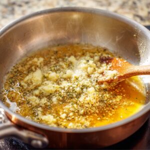 A saucepan filled with melted butter, minced garlic, Parmesan, and herbs simmering together, being stirred with a wooden spoon to make garlic butter glaze.