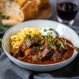 A bowl of German goulash served with buttered egg noodles, garnished with chopped parsley and a dollop of sour cream, with bread and red wine in the background.