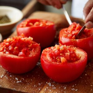 Close-up of hands preparing fresh tomatoes by scooping and filling them with a diced tomato mixture on a wooden cutting board.
