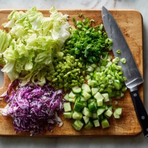 Chopped vegetables for green goddess salad on a wooden board — lettuce, cucumber, edamame, green onions, and shredded cabbage with a chef’s knife