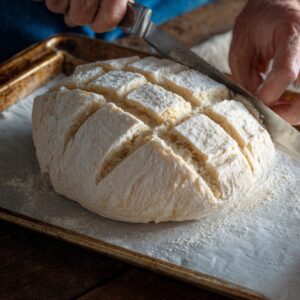 Hands scoring a round loaf of bread dough with a knife on a floured baking tray, preparing it for baking.