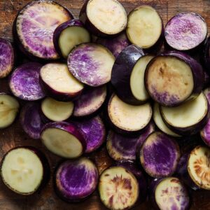 Easy Pasta Alla Norma Recipe 16 A pile of freshly sliced eggplant rounds showing vibrant purple skin and creamy white flesh, arranged on a wooden cutting board.