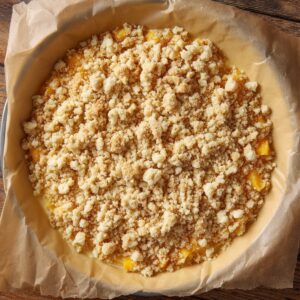 Unbaked peach crumble cheesecake in a springform pan lined with parchment, topped with a thick layer of golden crumble.
