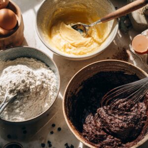 The Best Peanut Butter Brownie Swirl Cookies Recipe 15 A baking setup with bowls containing brownie batter, peanut-butter dough, and dry ingredients. Broken eggshells, chocolate chips, and whisks are scattered around the workspace in natural sunlight.