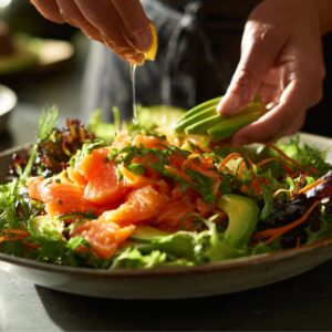 The Best Smoked Salmon Salad Recipe 16 A person squeezes fresh lemon juice over a colorful salad topped with slices of smoked salmon, avocado, shredded carrots, and mixed greens in warm natural light.