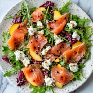 The Best Smoked Salmon Salad Recipe 17 A vibrant smoked salmon salad with mixed greens, peaches, goat cheese, and crushed pepper, arranged beautifully on a speckled ceramic plate.